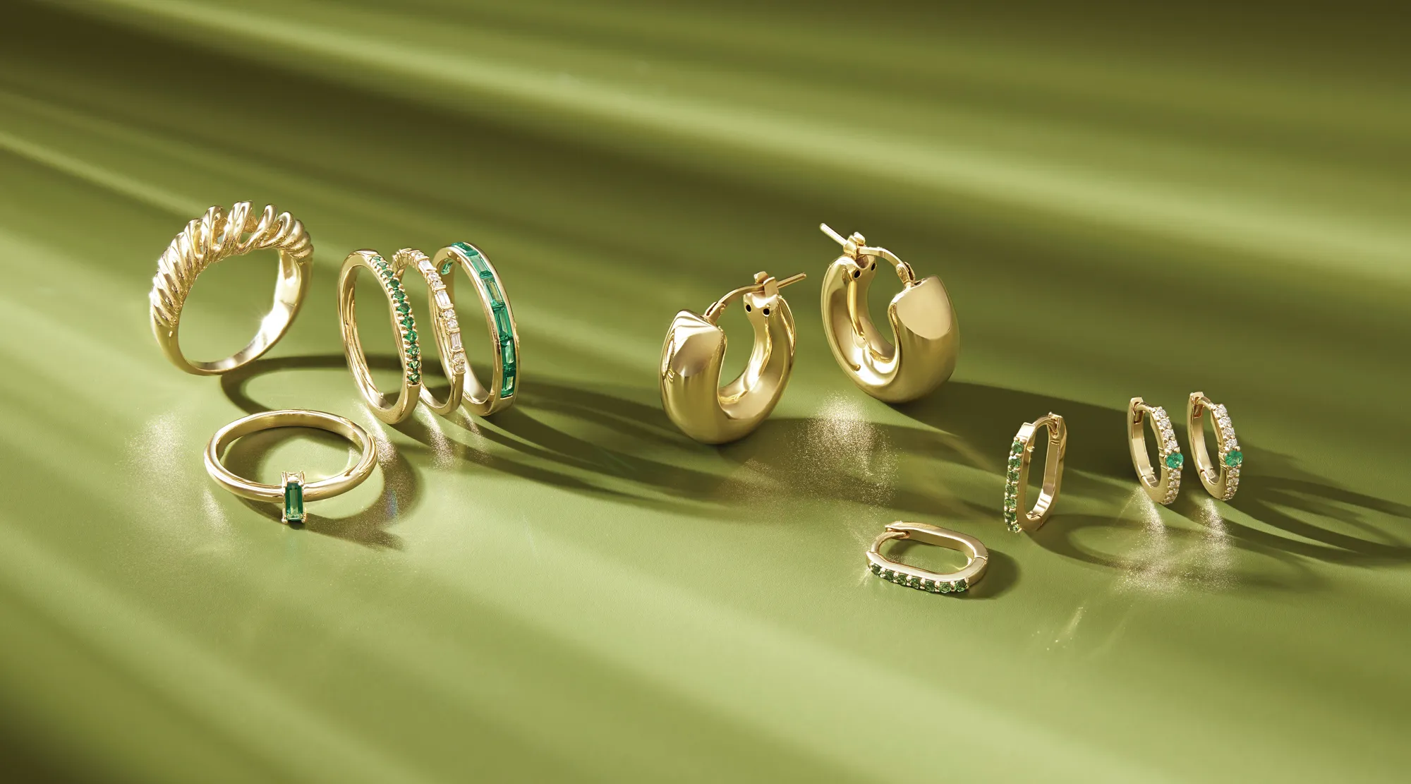 Gold earrings and rings with emerald accents displayed on a green background. 