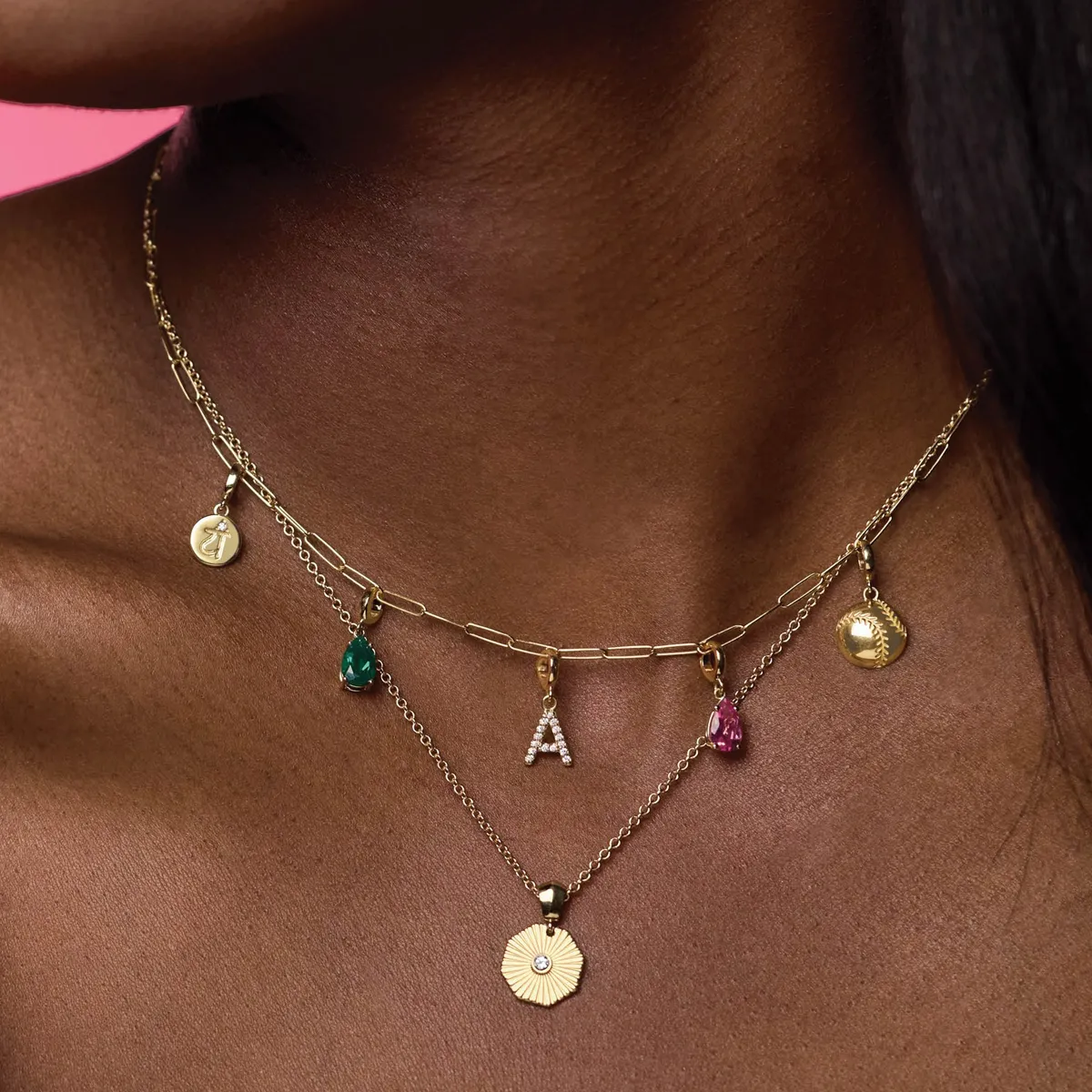Gold layered necklace with colorful charms, including an 'A', emerald, and baseball pendant.