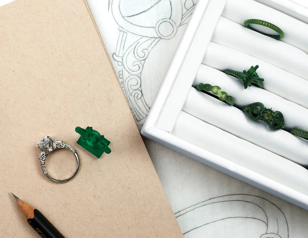 Custom engagement rings displayed with a diamond ring and green wax models on a sketch background.