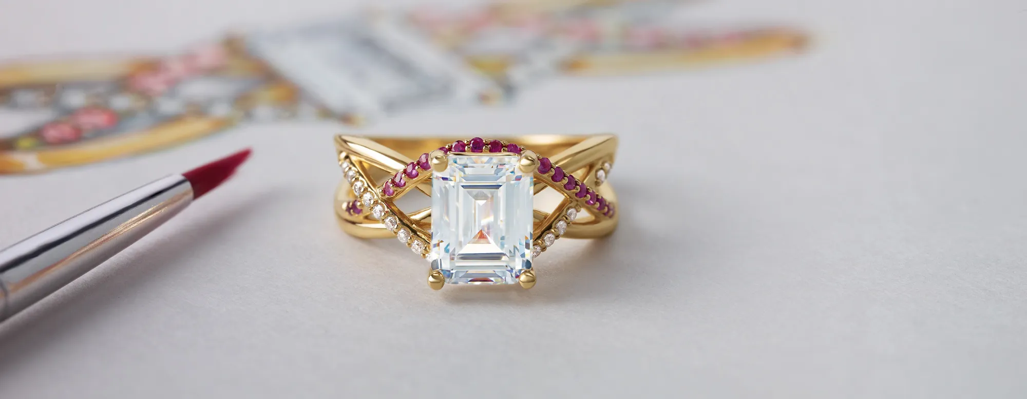 Elegant gold ring with a large emerald-cut diamond and ruby accents, alongside a paintbrush on a design sketch.