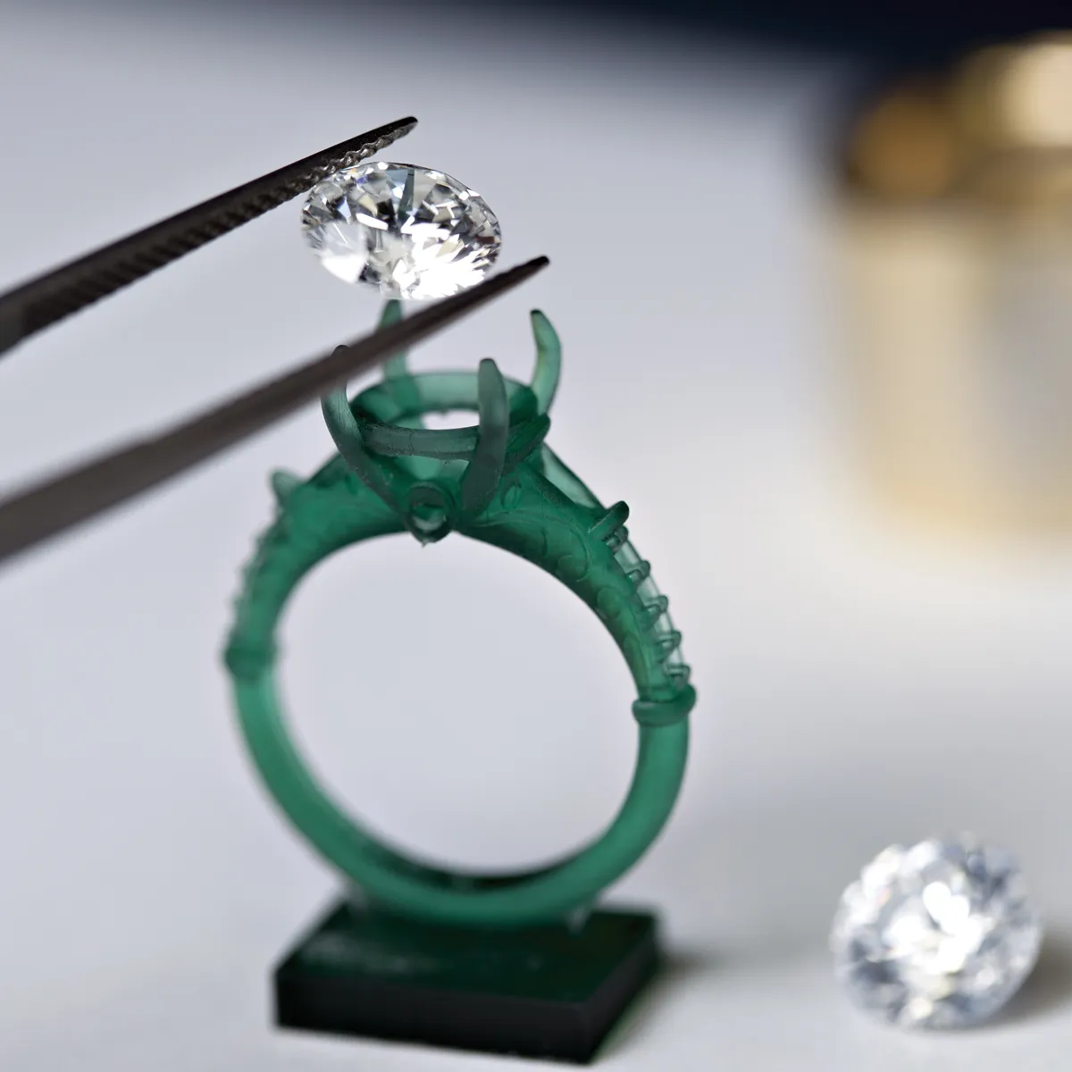 A jeweler using tweezers to place a diamond on a green ring setting, with another diamond nearby.