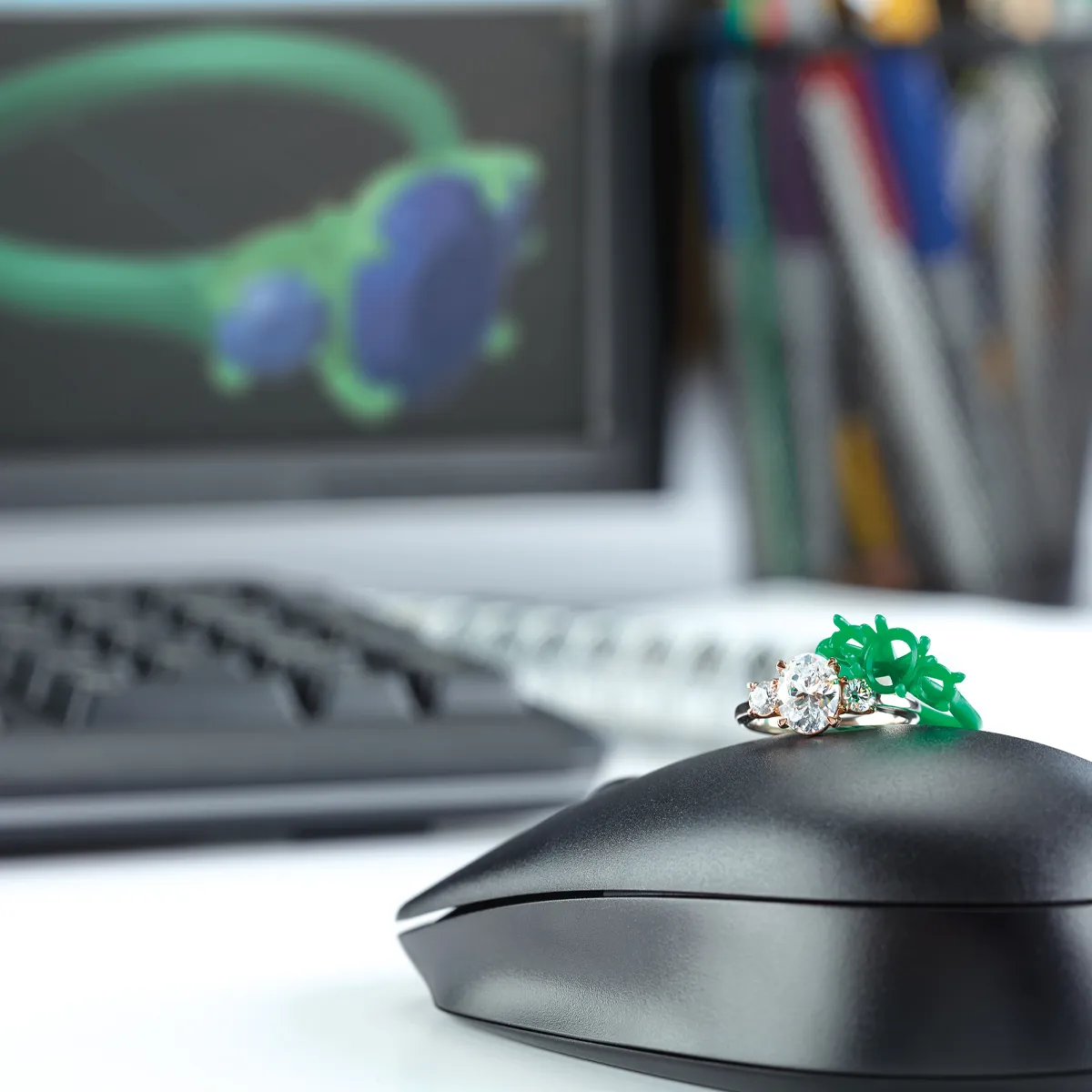 A diamond engagement ring and a green ring sizer on a computer mouse, with a blurred 3D ring design on the screen behind.