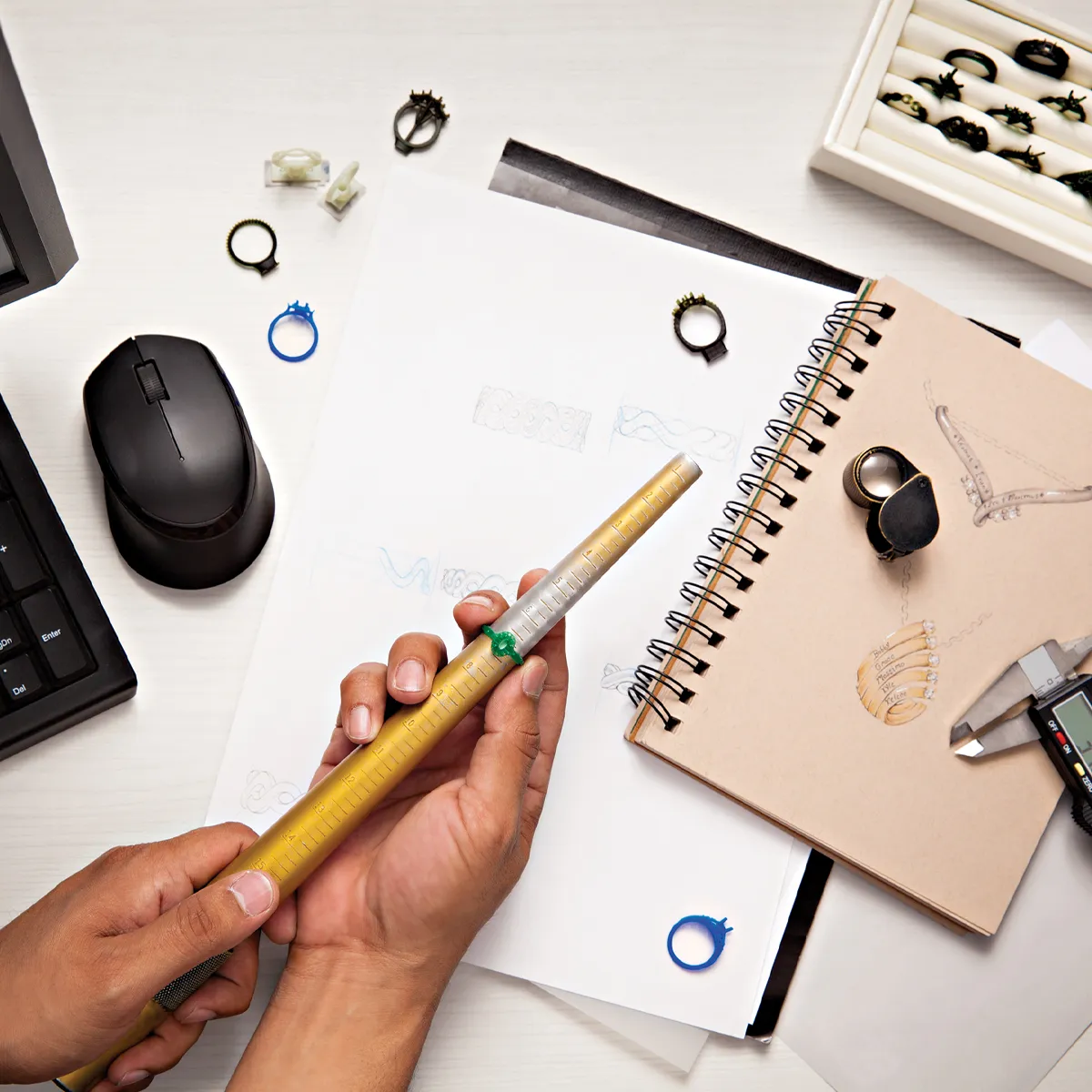 A hand holding a ring sizer with sketches and jewelry tools on a desk, showcasing custom jewelry design work.