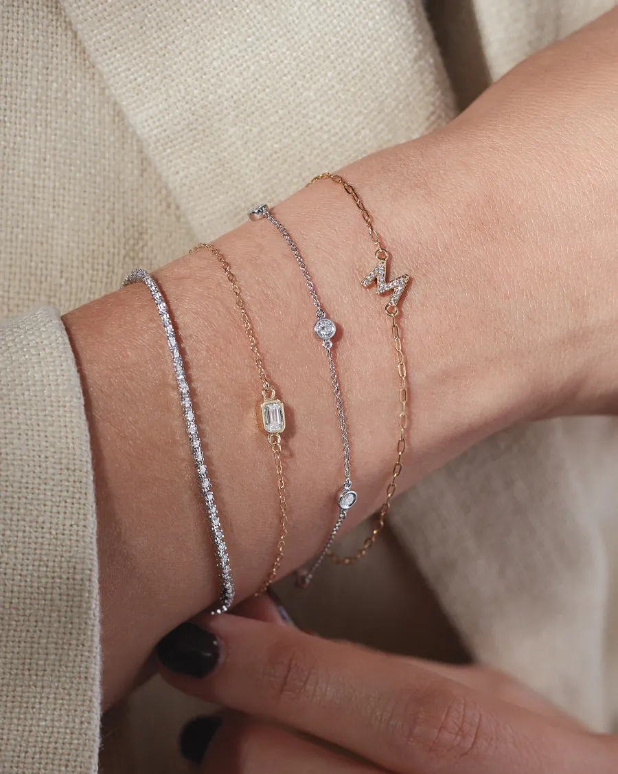 Four elegant bracelets in gold and silver tones, featuring diamonds and a letter charm, worn on a wrist.