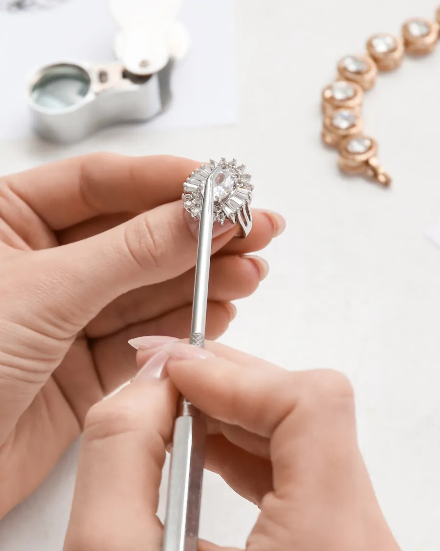 A person inspecting a sparkling silver ring with a tool, surrounded by jewelry pieces on a table.