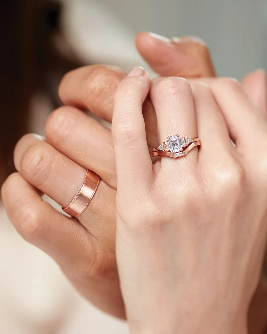 Two hands wearing rose gold rings, one with an emerald-cut diamond engagement ring, showcasing love and commitment.
