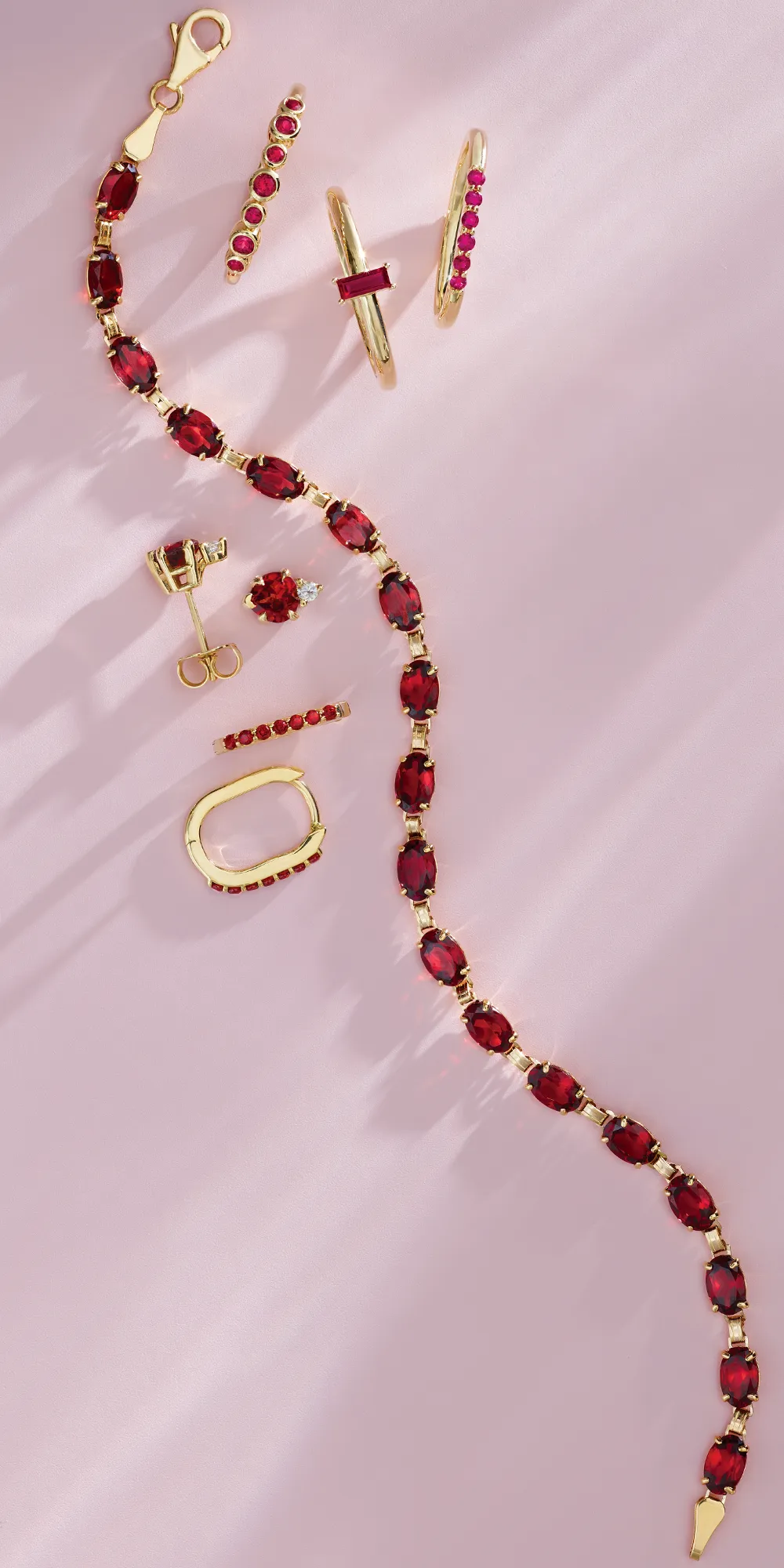 Elegant gold jewelry featuring red gemstones, including rings, earrings, and a bracelet, displayed on a soft pink background.