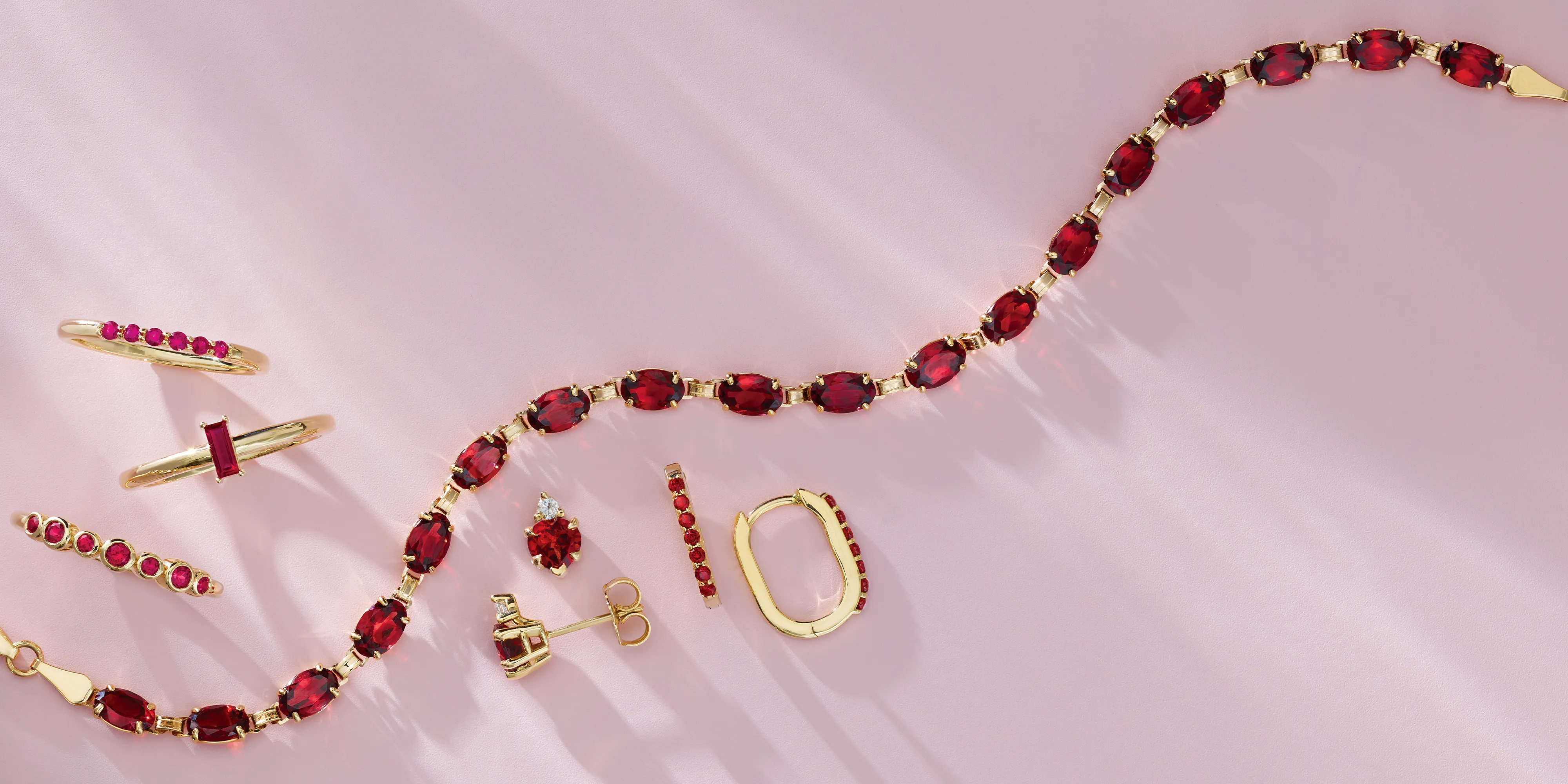 Elegant gold jewelry featuring red gemstones, including rings, earrings, and a bracelet, displayed on a soft pink background.