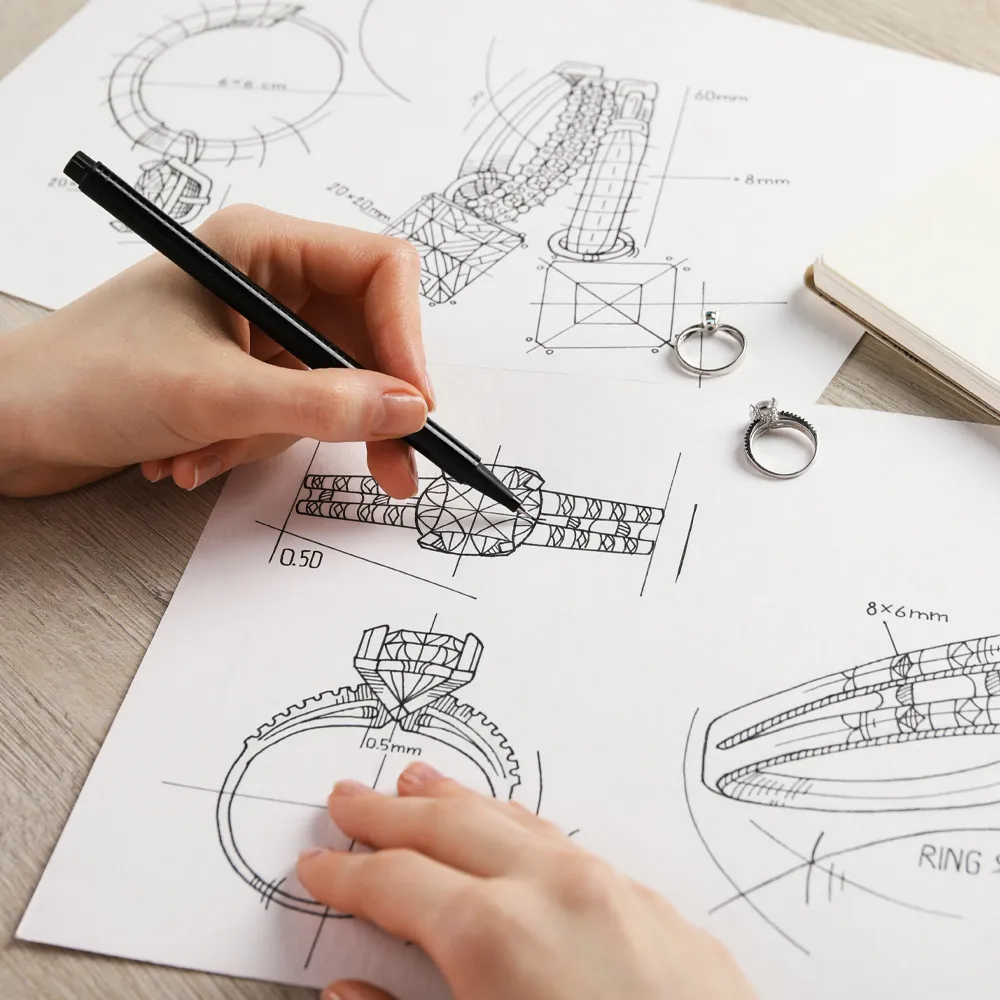 A hand sketches jewelry designs on paper, featuring rings and a bracelet, with two rings placed beside the drawings.