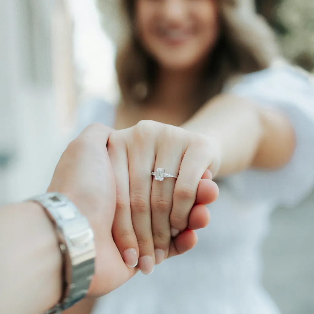 A couple holding hands, showcasing an engagement ring, with a blurred background of a sunny street.