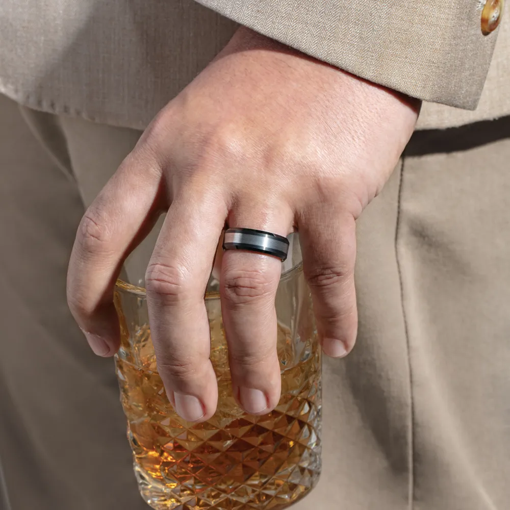 A hand holding a glass of whiskey, wearing a black and silver wedding band, against a beige suit background.