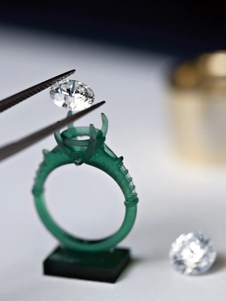 A jeweler using tweezers to place a diamond on a green ring setting, with another diamond nearby.