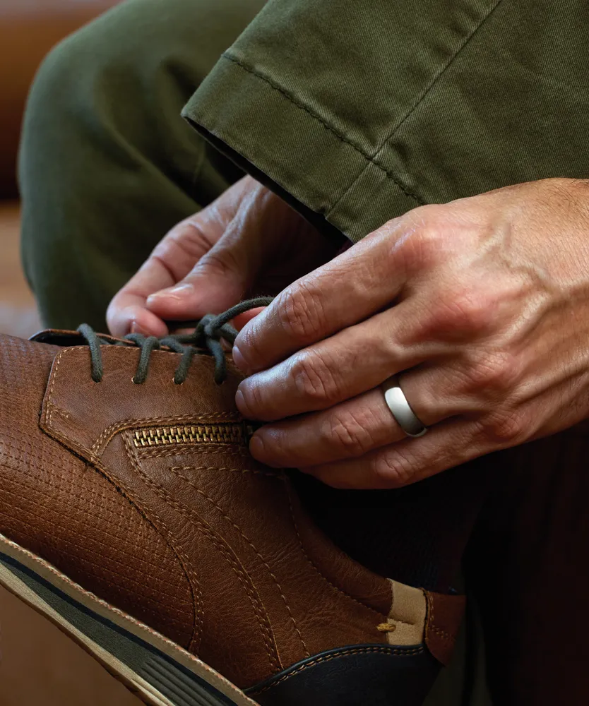 A person tying brown leather shoes while wearing a wedding band, seated with green pants.