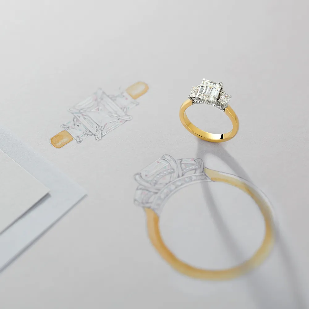 A gold engagement ring with a large diamond sits beside its sketch on paper.