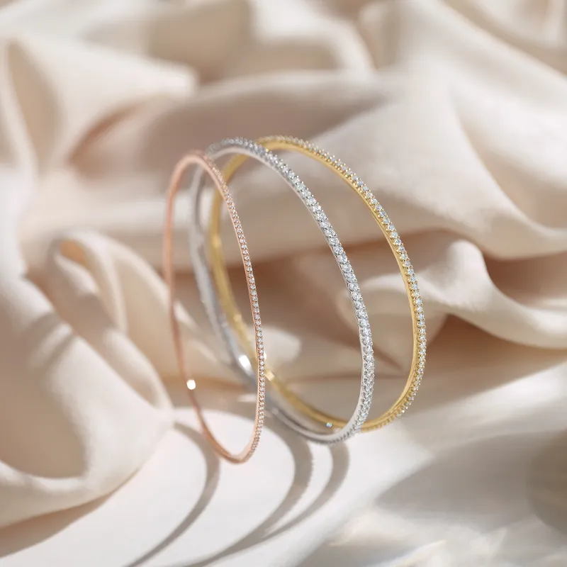 Three elegant bangles in rose gold, silver, and yellow gold, adorned with sparkling stones, resting on soft fabric.