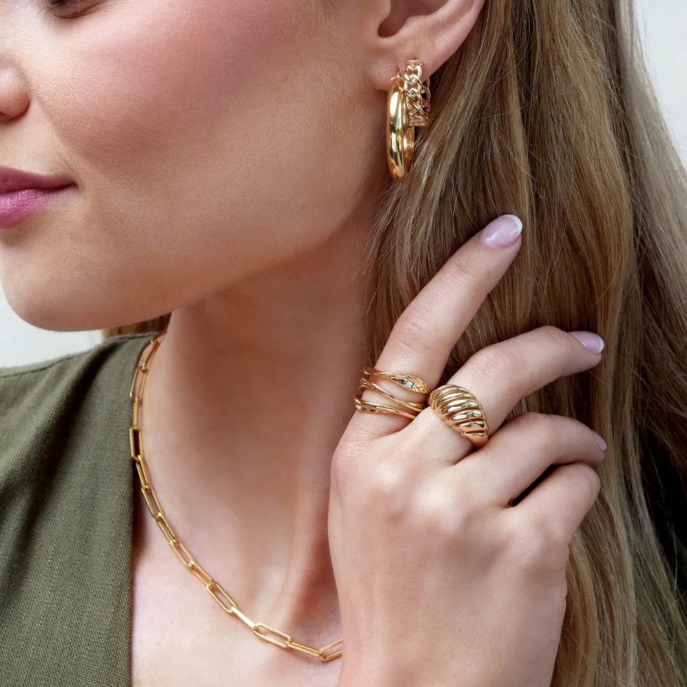 Close-up of a woman wearing gold jewelry: hoop earrings, layered rings, and a chain necklace. Shop at Ambassador Diamond Jeweler