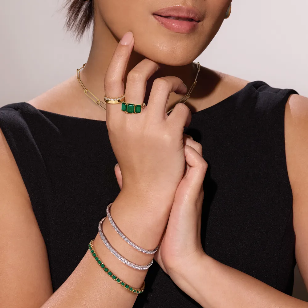Model showcasing elegant jewelry: green gemstone ring, bracelets, and gold chain necklace against a neutral background.