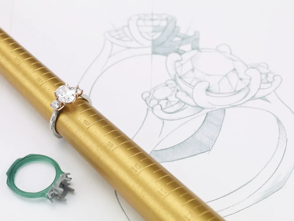 A diamond engagement ring next to a design sketch on a gold ring mandrel, showcasing jewelry craftsmanship.