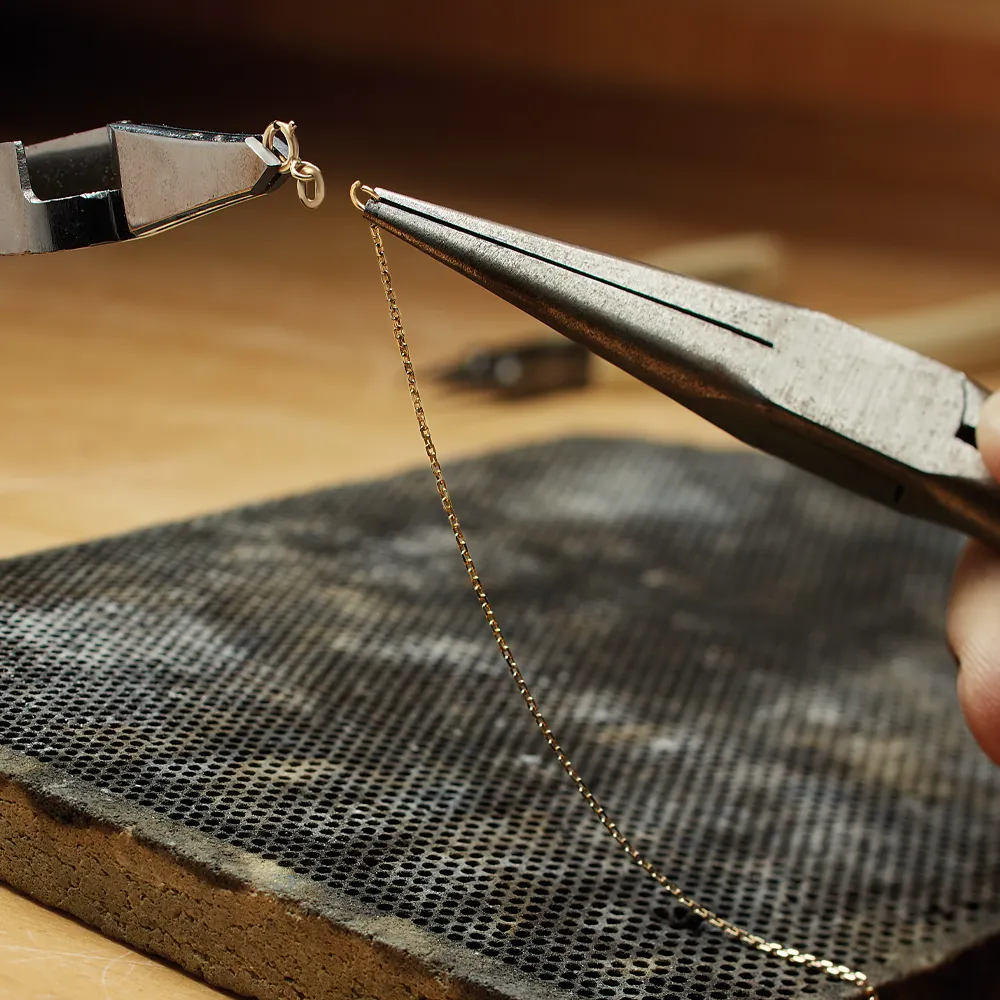 A jeweler using pliers to attach a gold chain link, with a textured work surface in the background.