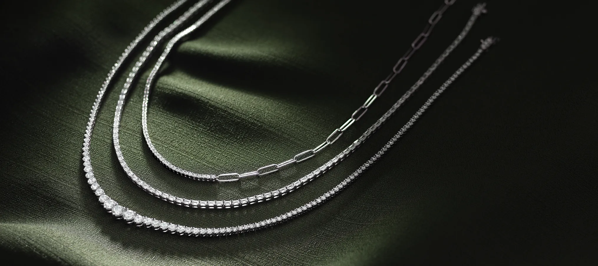 Three elegant silver necklaces displayed on a dark green fabric background. Shop at Amos Jewelry for fine jewelry.