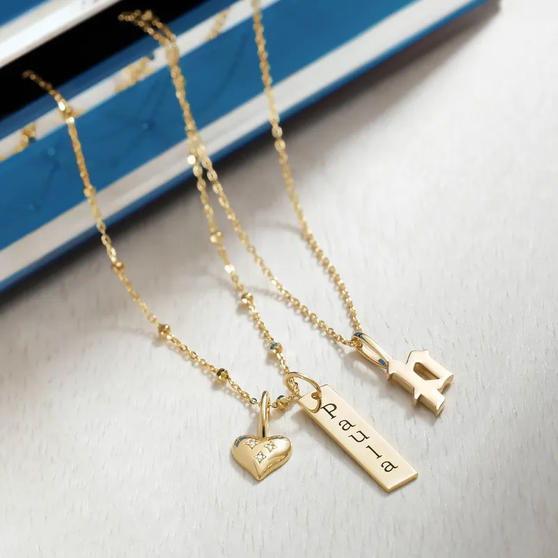 Three gold necklaces with charms: a heart, a nameplate, and a letter, displayed on a light surface.
