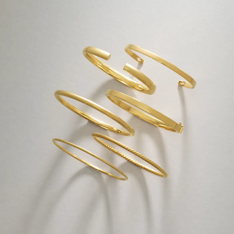 Five elegant gold bangles of varying styles arranged on a light background. Shop at Amos Jewelry in Wray.
