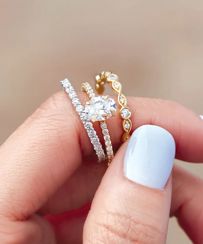 A hand holding three elegant rings, featuring diamonds and gold, with a light blue manicure. 