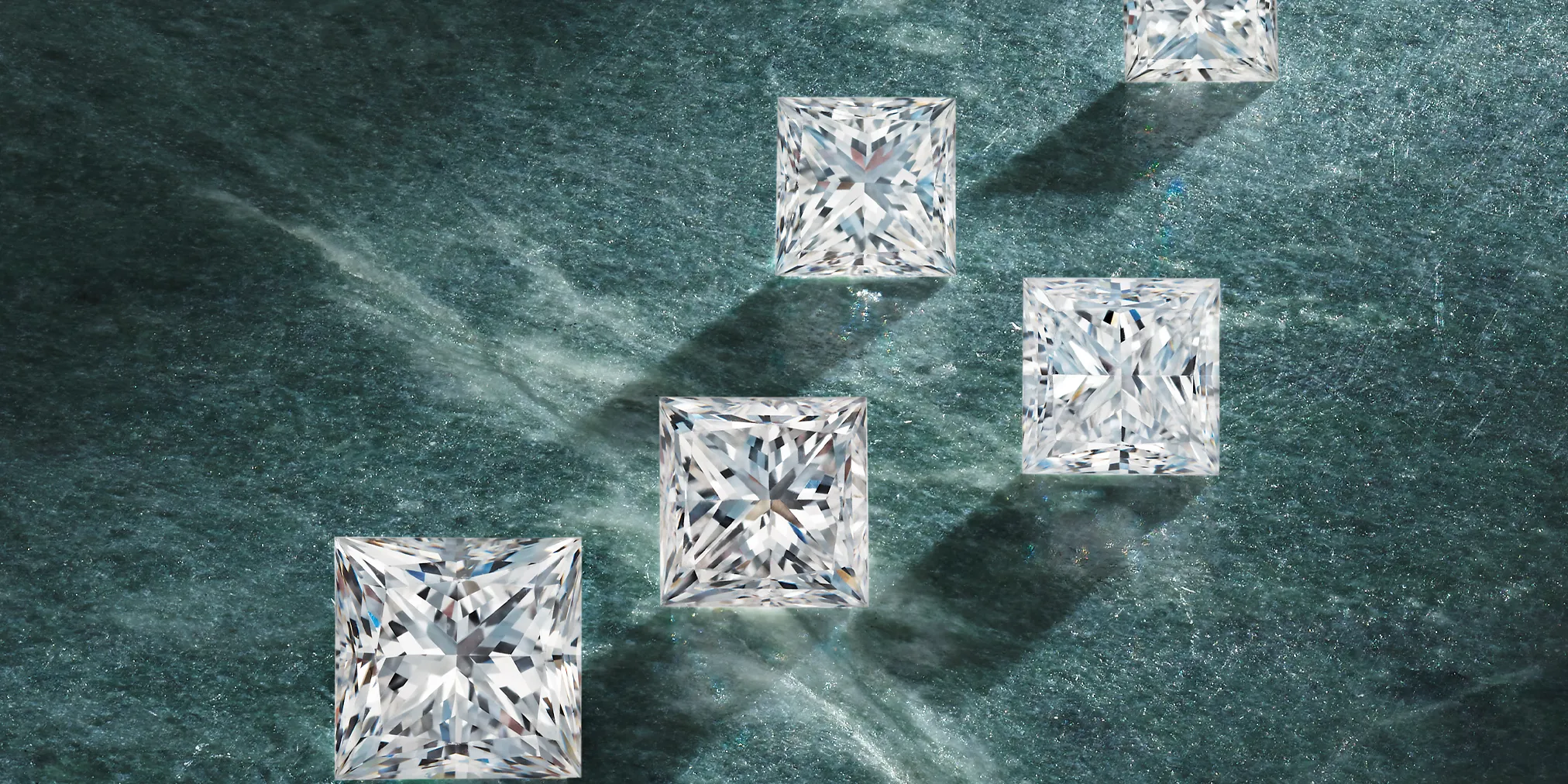 Four sparkling rectangular diamonds casting shadows on a dark green surface. Click to make an appointment at Amos Jewelry.