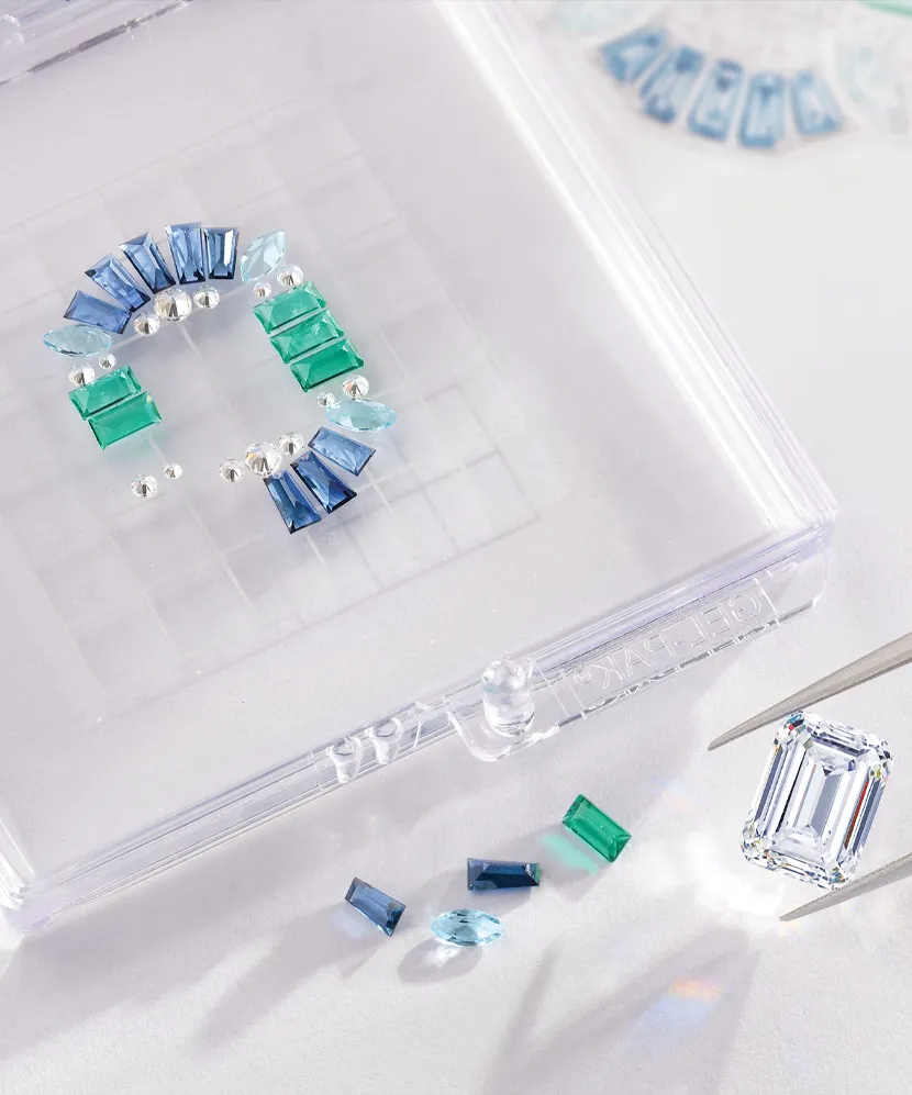 Colorful gemstones arranged in a display, with a large emerald-cut stone being held by tweezers. Visit Amos Jewelry for more.