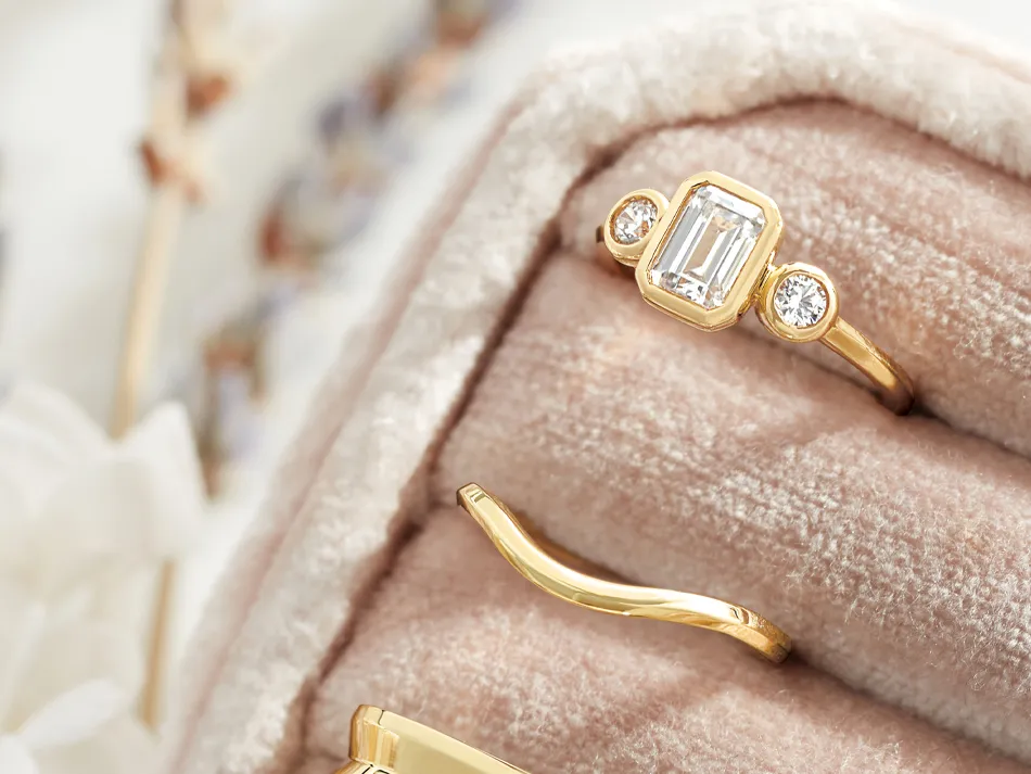 Elegant gold engagement ring with an emerald-cut diamond and two round diamonds, displayed on a plush ring holder.