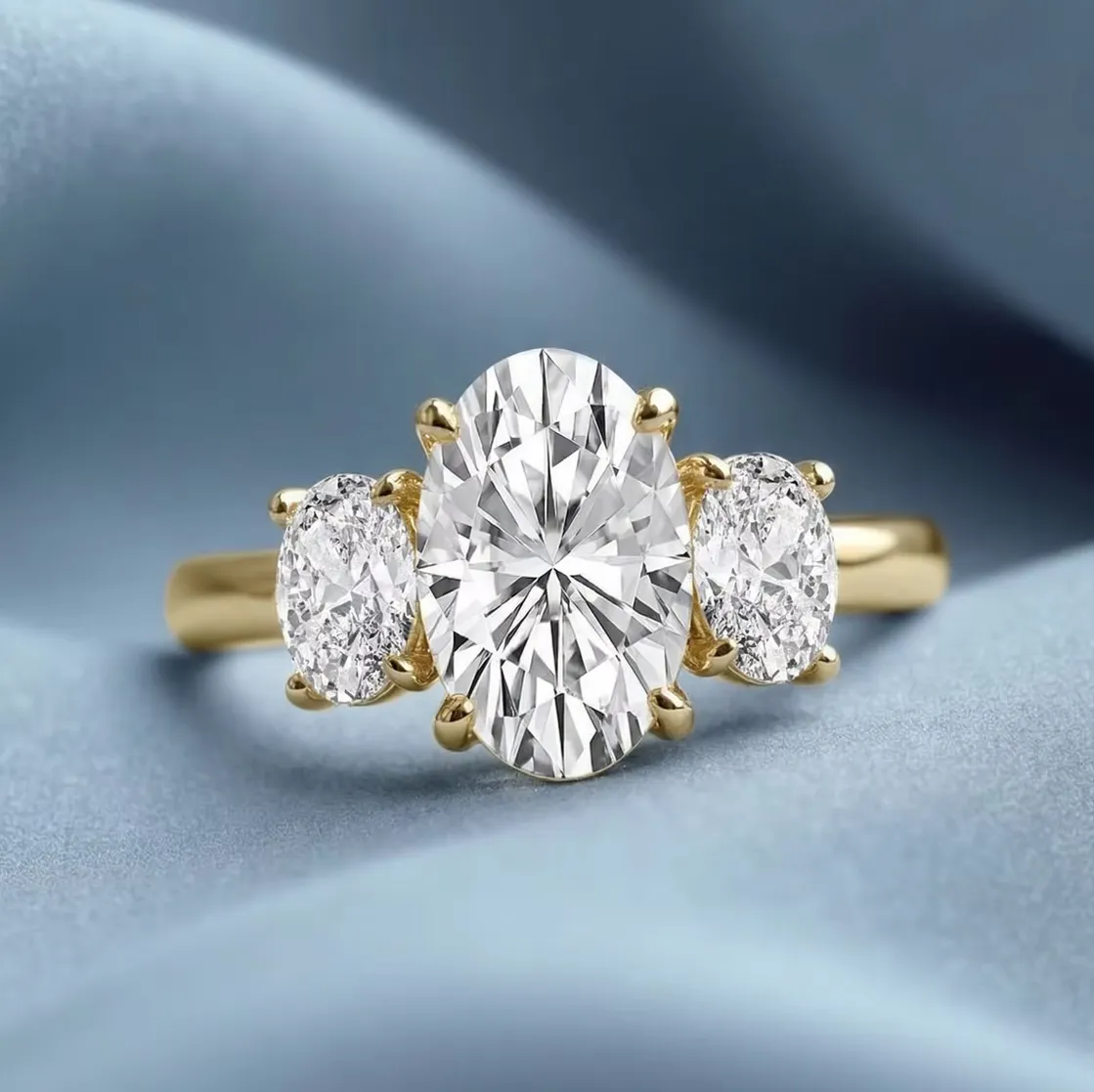 A gold ring with a large oval diamond center stone and two smaller side diamonds on a blue fabric background.