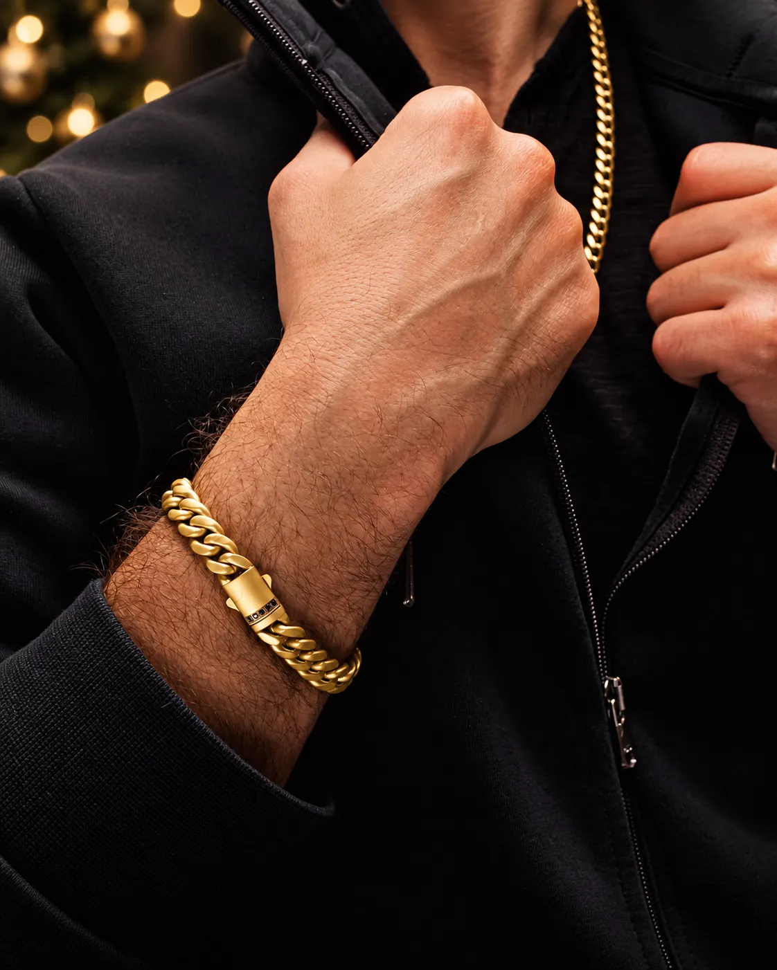 Man wearing a gold-tone chain and curb-link bracelet with black clothing and warm holiday lights