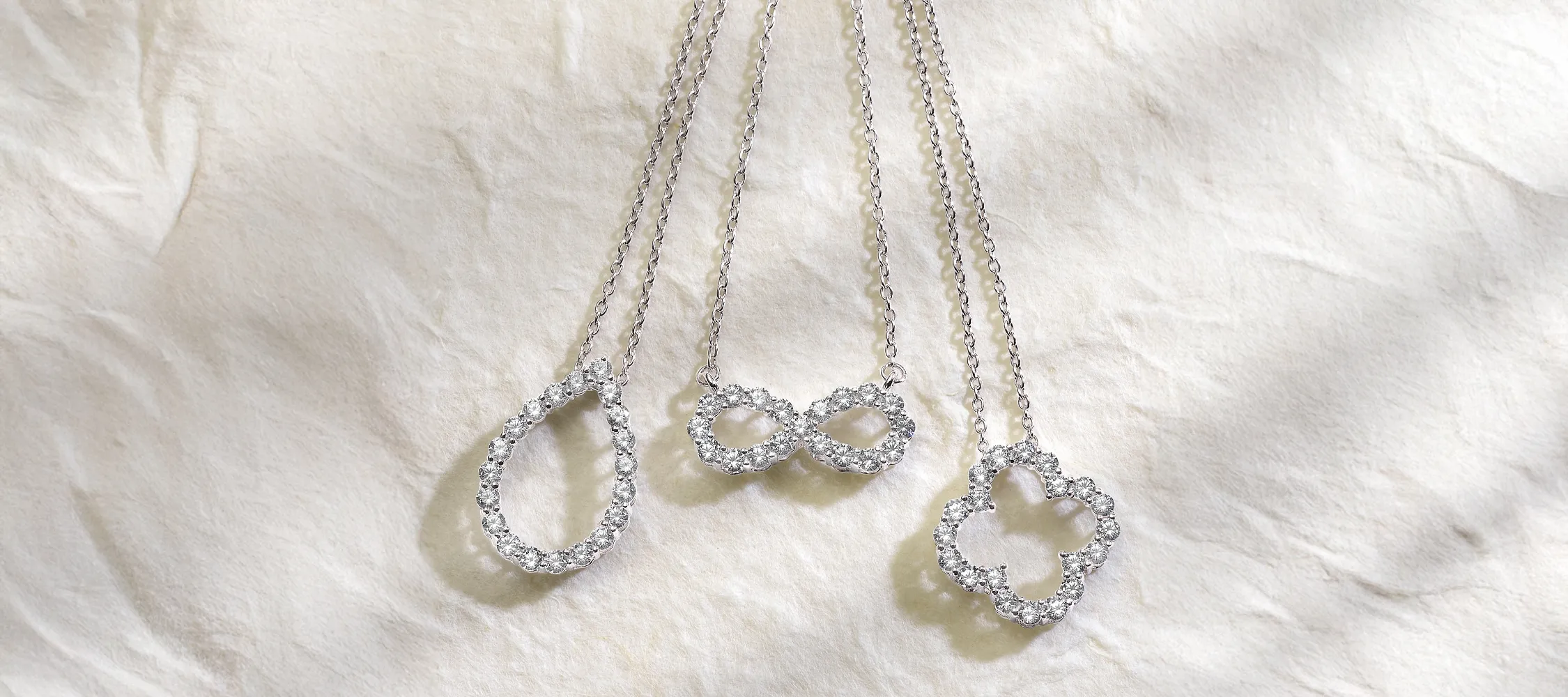 Three elegant silver necklaces with sparkling pendants: teardrop, infinity, and floral shapes on a soft background.