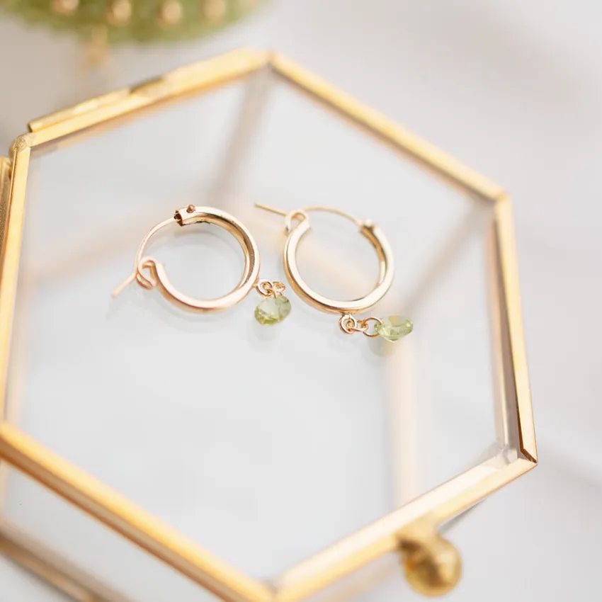 Gold hoop earrings with green gemstone charms displayed in a glass case.