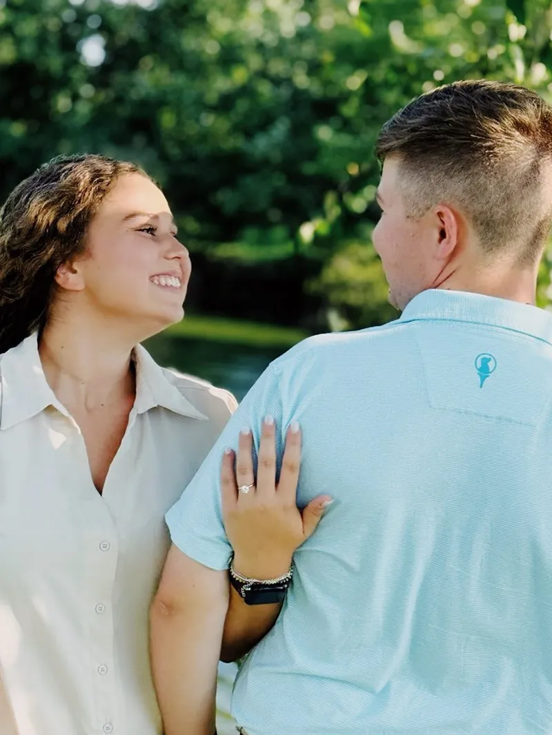 Couple smiling at each other, showcasing an engagement ring in a lush outdoor setting.