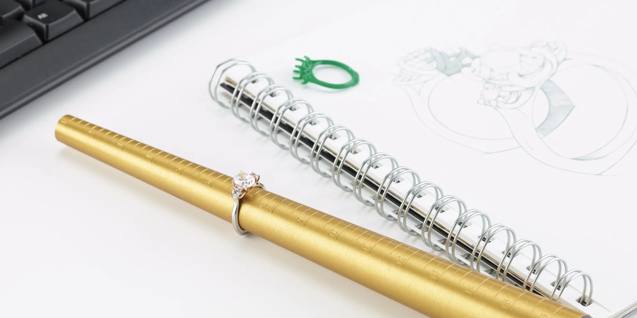A gold ring sizer with a diamond ring and a green ring mold on a desk with a sketchbook and keyboard.