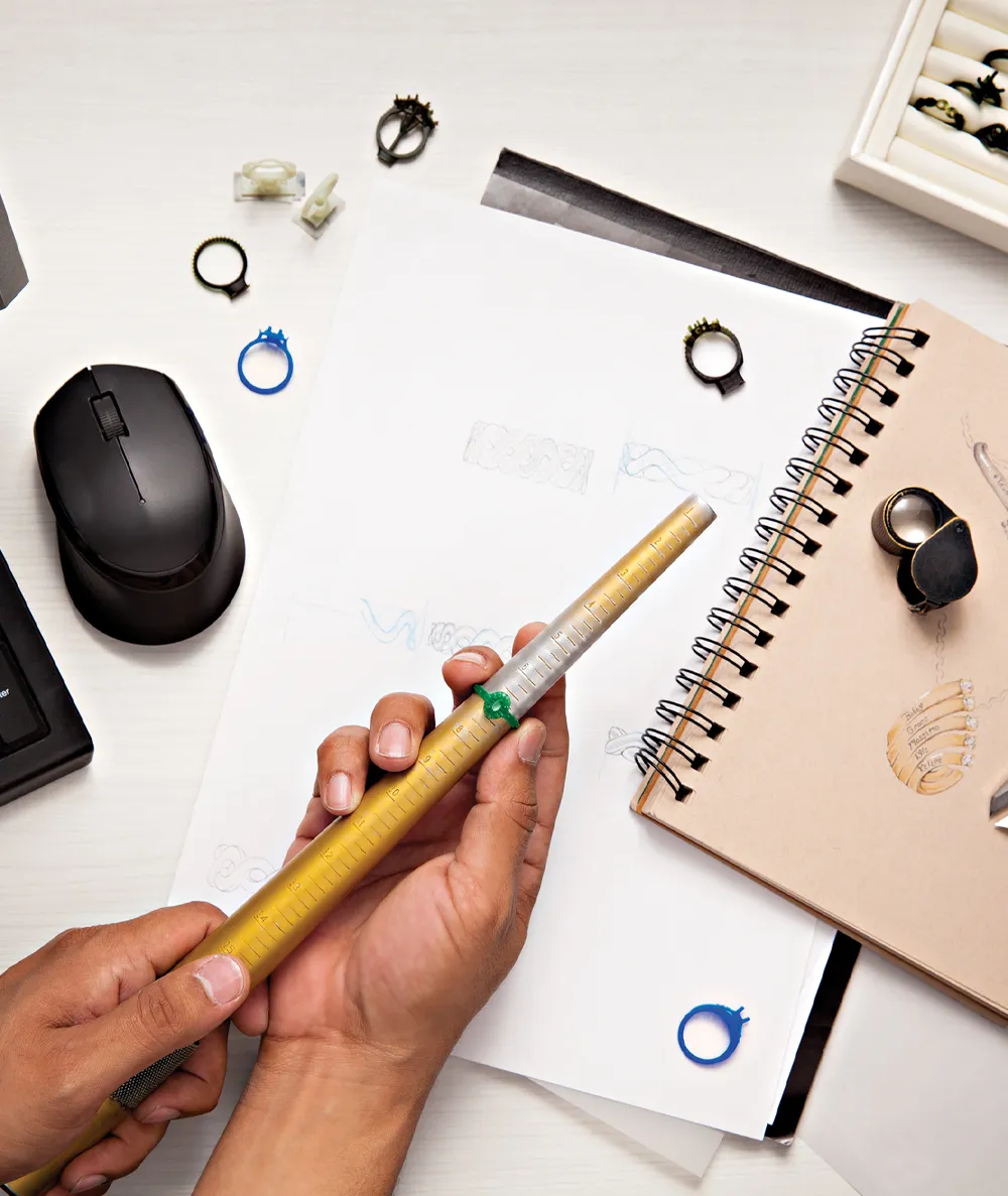A hand holding a ring sizer with various rings and sketches on a desk, showcasing jewelry design work.