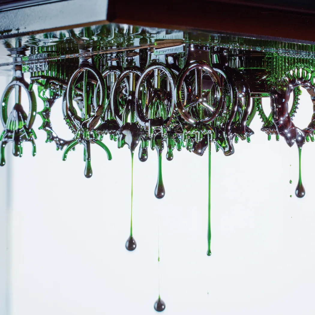 Dripping green liquid forms intricate shapes resembling jewelry, suspended from a reflective surface.