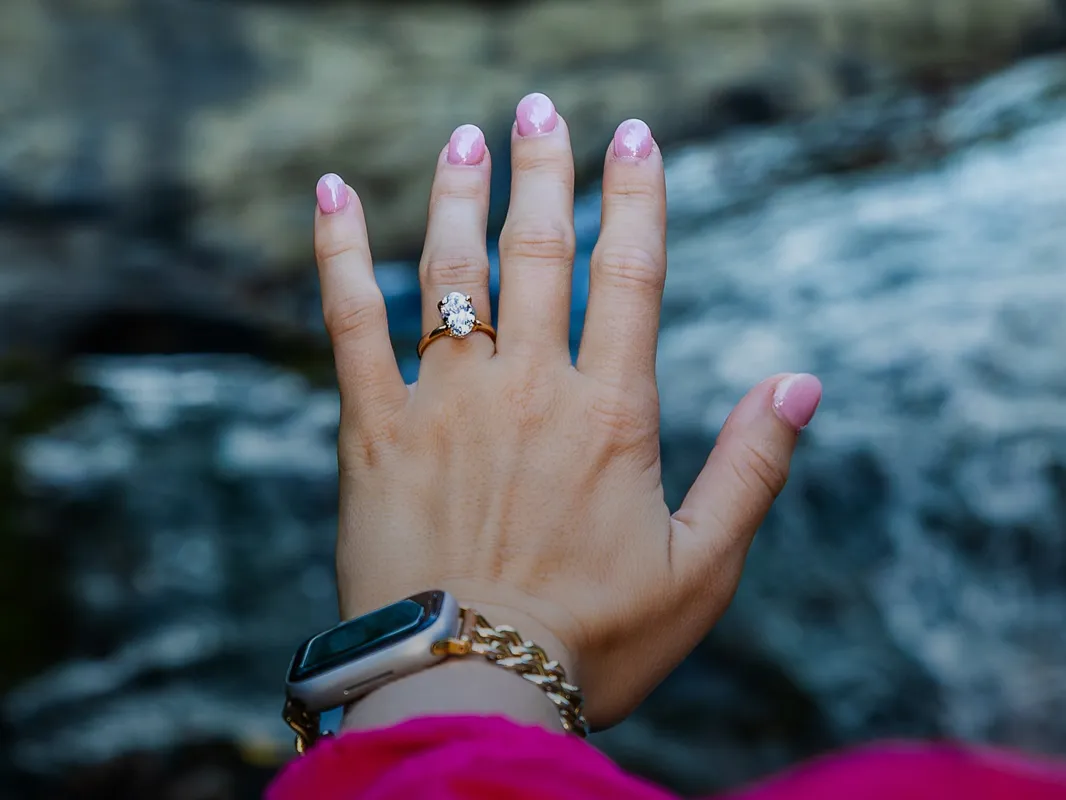 Hand displaying an engagement ring with a diamond, pink nails, and a smartwatch, set against a blurred natural background.