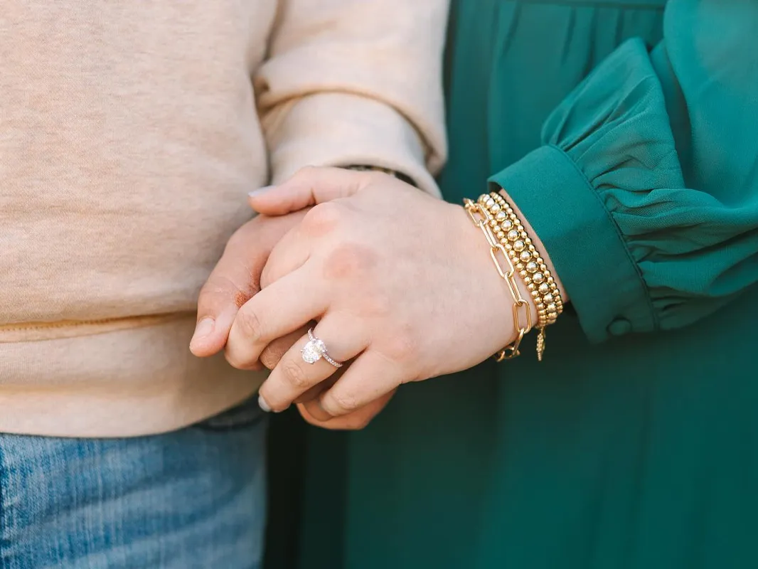 A couple holding hands, showcasing an engagement ring and gold bracelets.