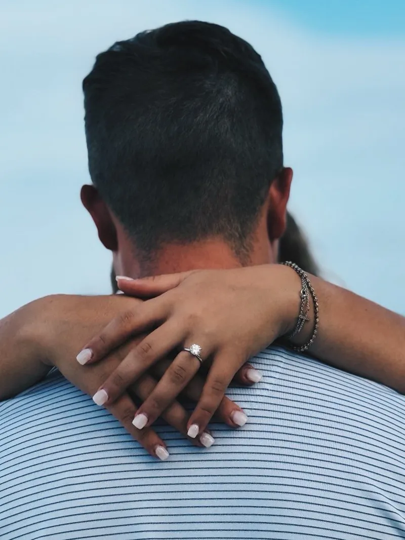 Couple embracing, showcasing an engagement ring against a blue background.