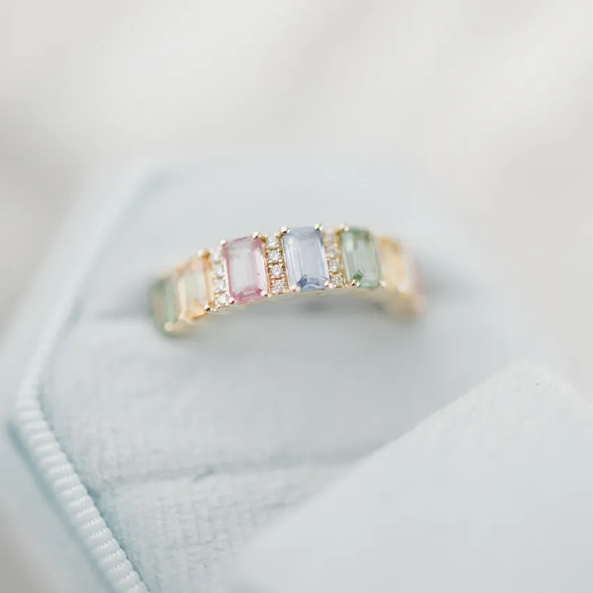 Colorful gemstone ring with pink, blue, and green stones set in gold, displayed on a soft blue velvet box.
