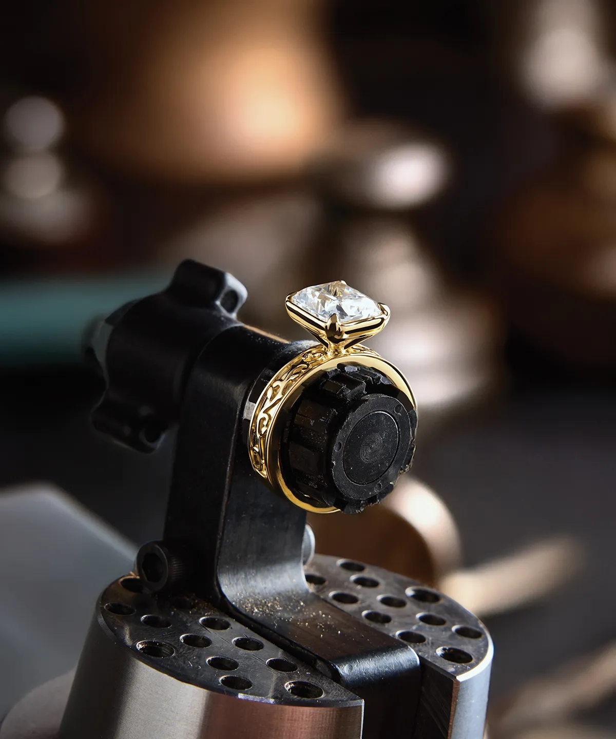 A gold ring with a large diamond set in a jeweler's clamp, blurred background.