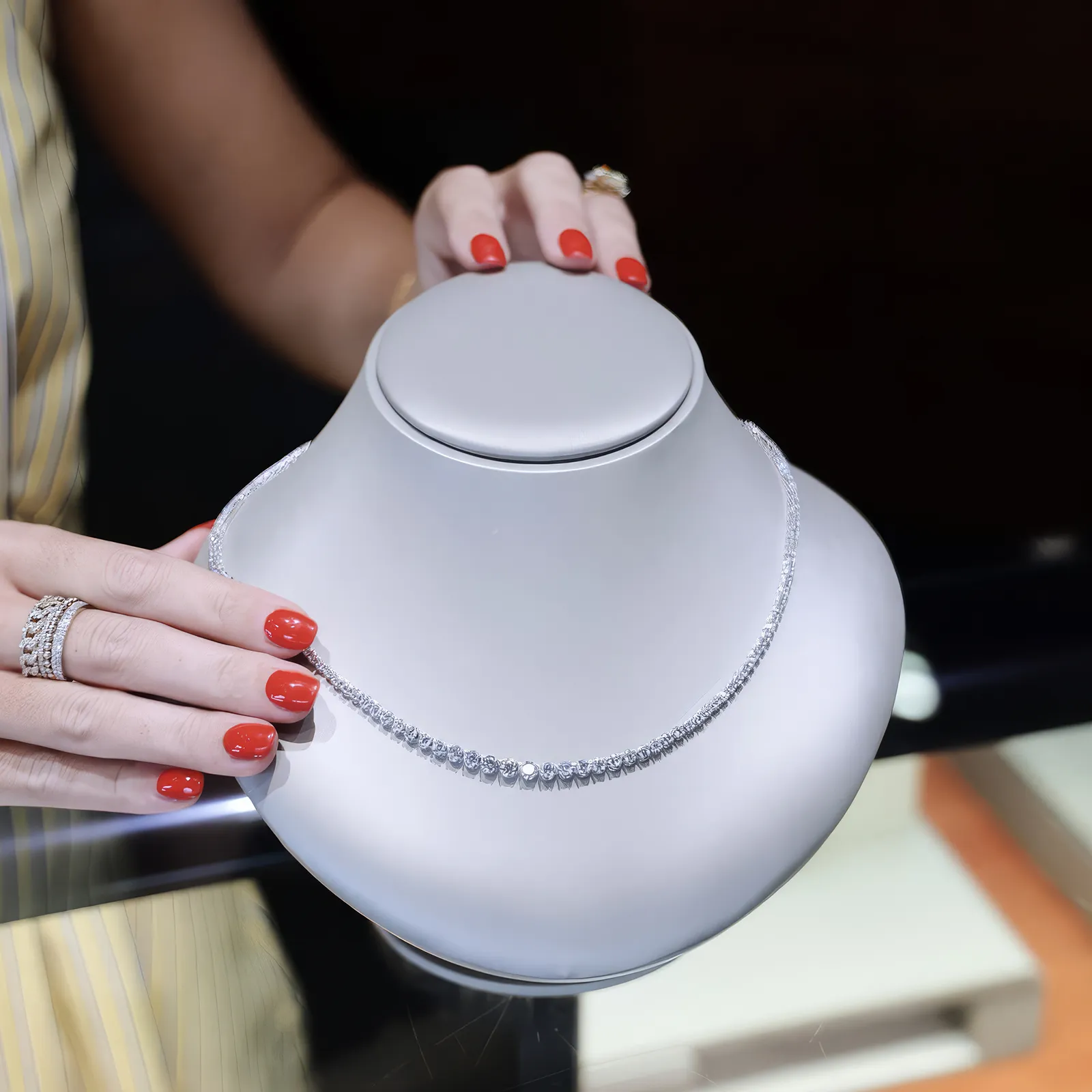 A person with red nails holds a diamond necklace on a display bust.