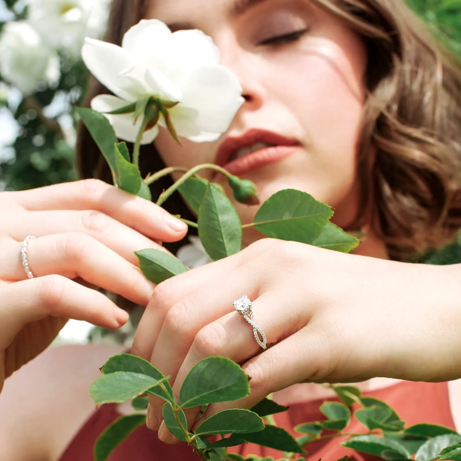 A woman with closed eyes holds a white flower, showcasing diamond rings on her fingers.