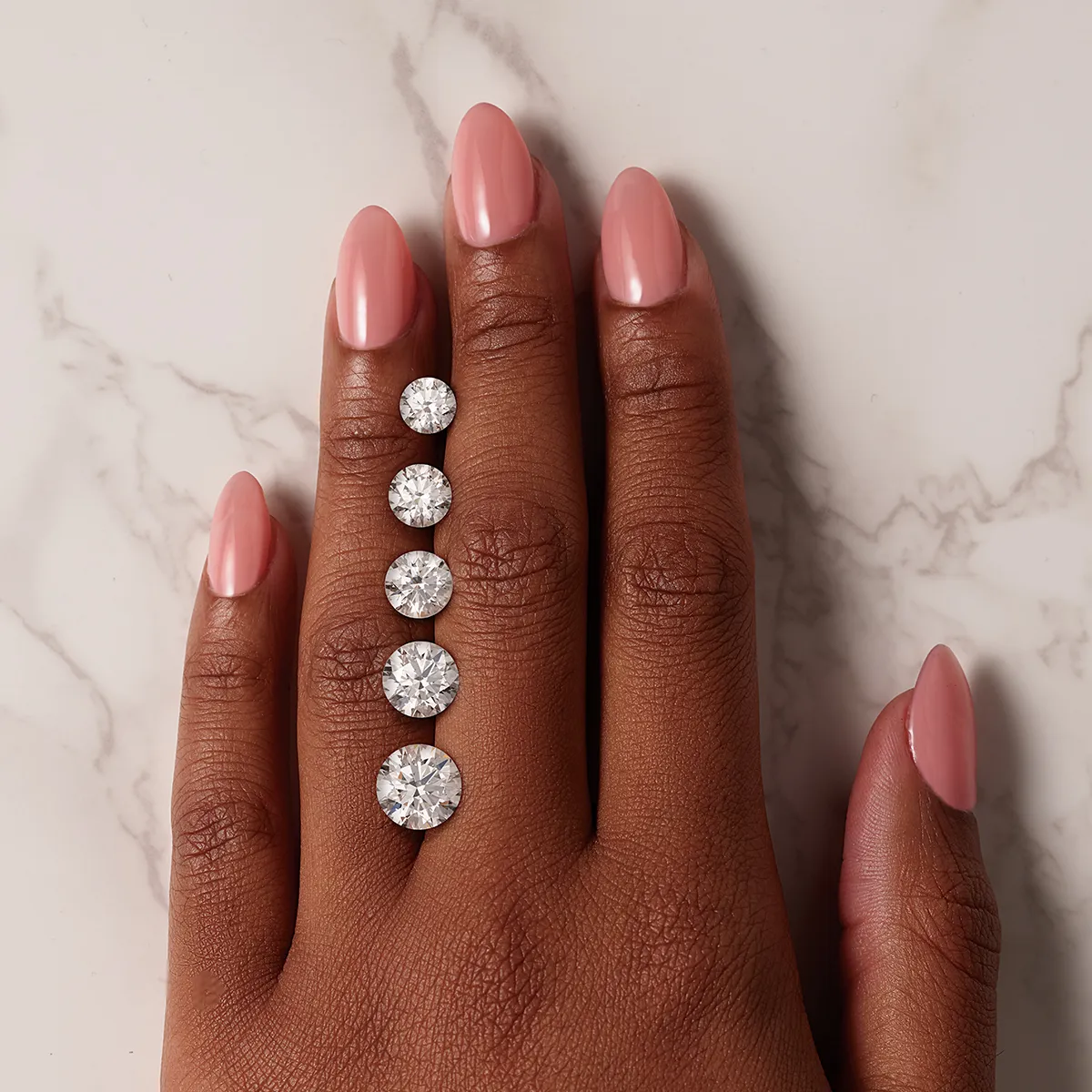 A hand with pink nails displays five loose diamonds of varying sizes on a marble surface.