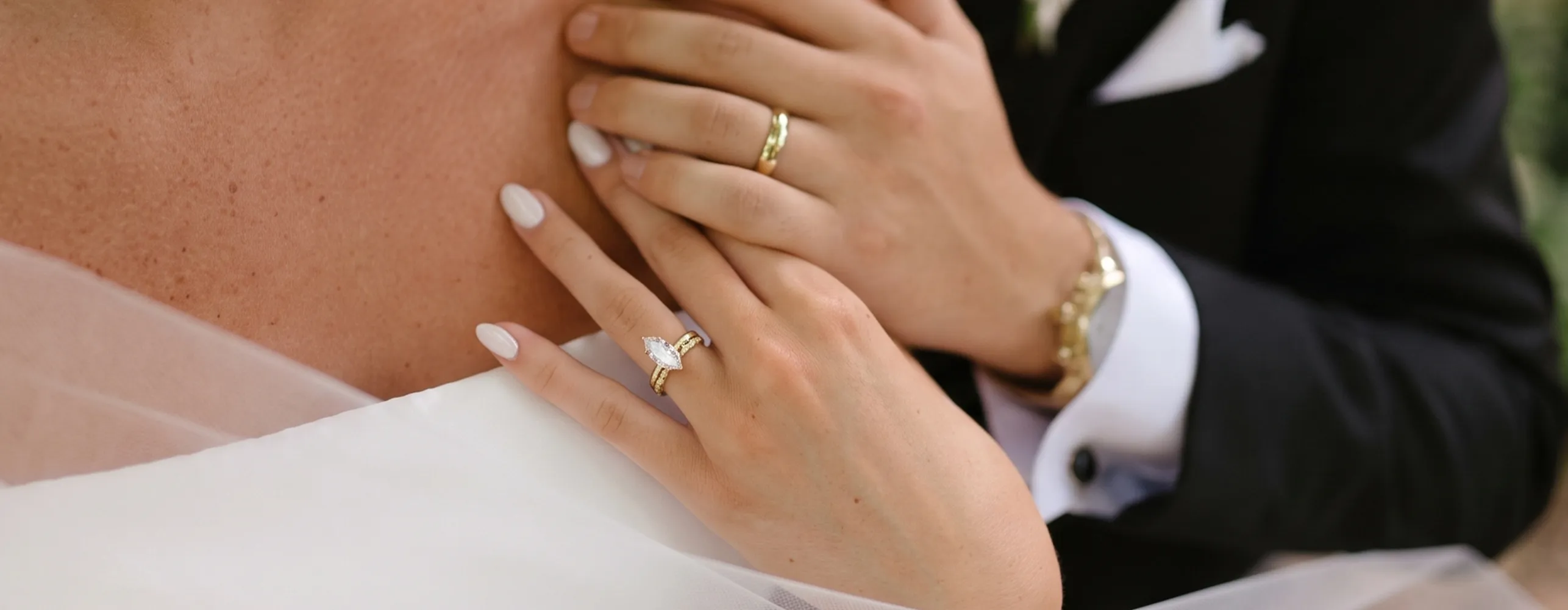 A close-up of hands with wedding rings, one featuring a marquise diamond, against a white dress.