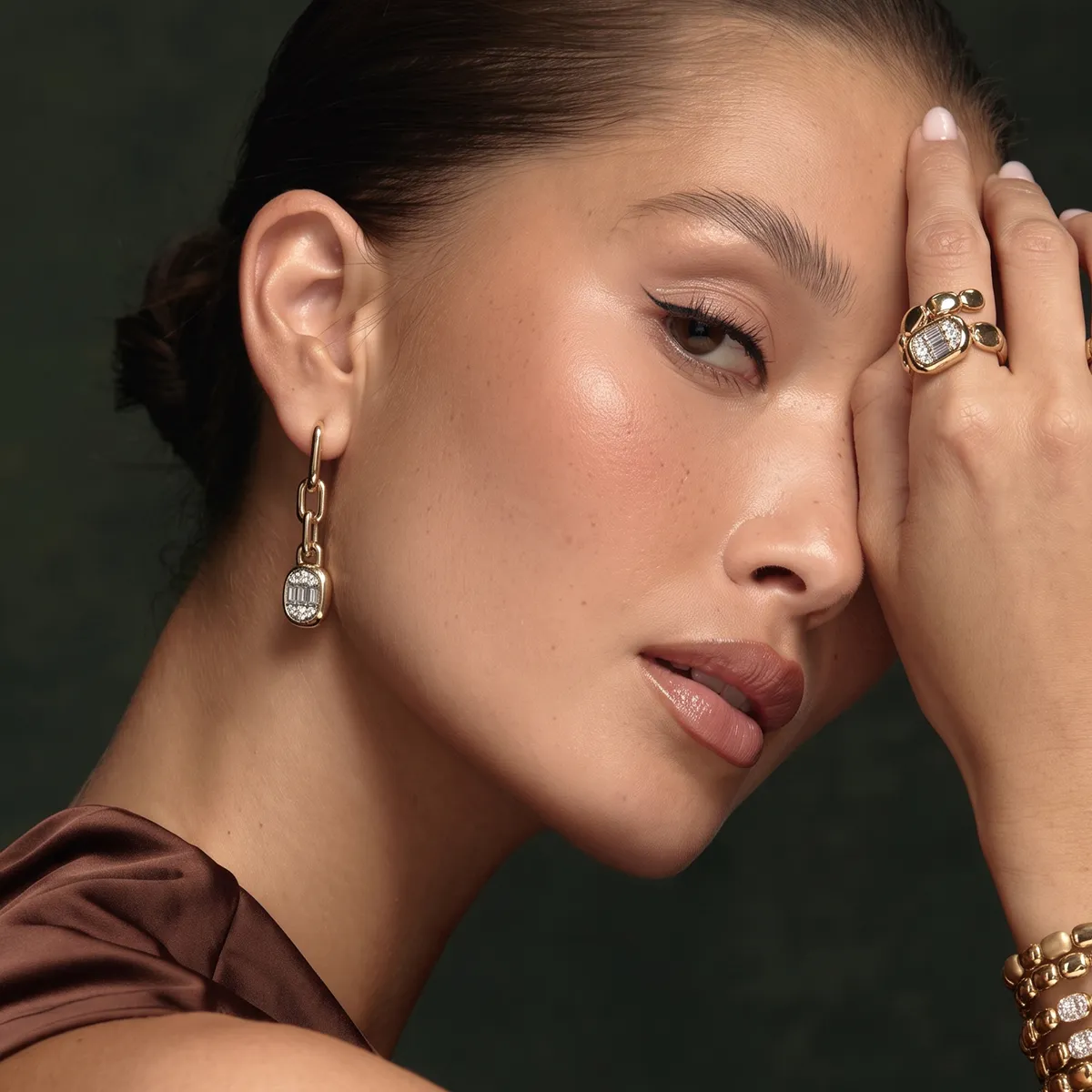 A woman wearing gold earrings and a ring, with her hand partially covering her face.