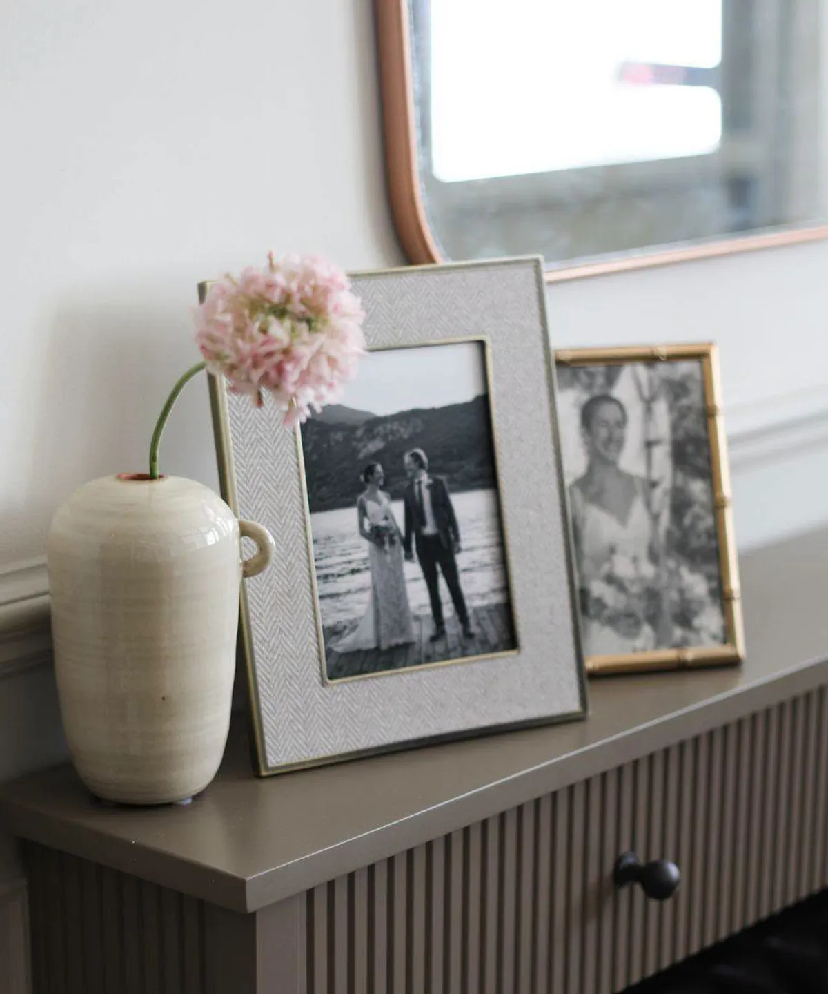 A vase with a pink flower and two framed photos on a table in front of a mirror.
