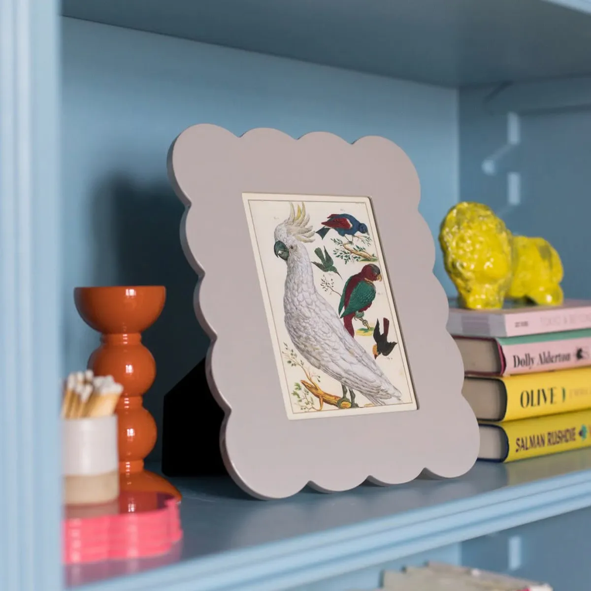 Shelf with framed bird illustration, orange candle holder, yellow lion figurine, and stacked books.